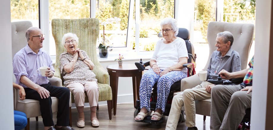Male And Female Residents Sitting In Chairs And Talking In Lounge Of Retirement Home