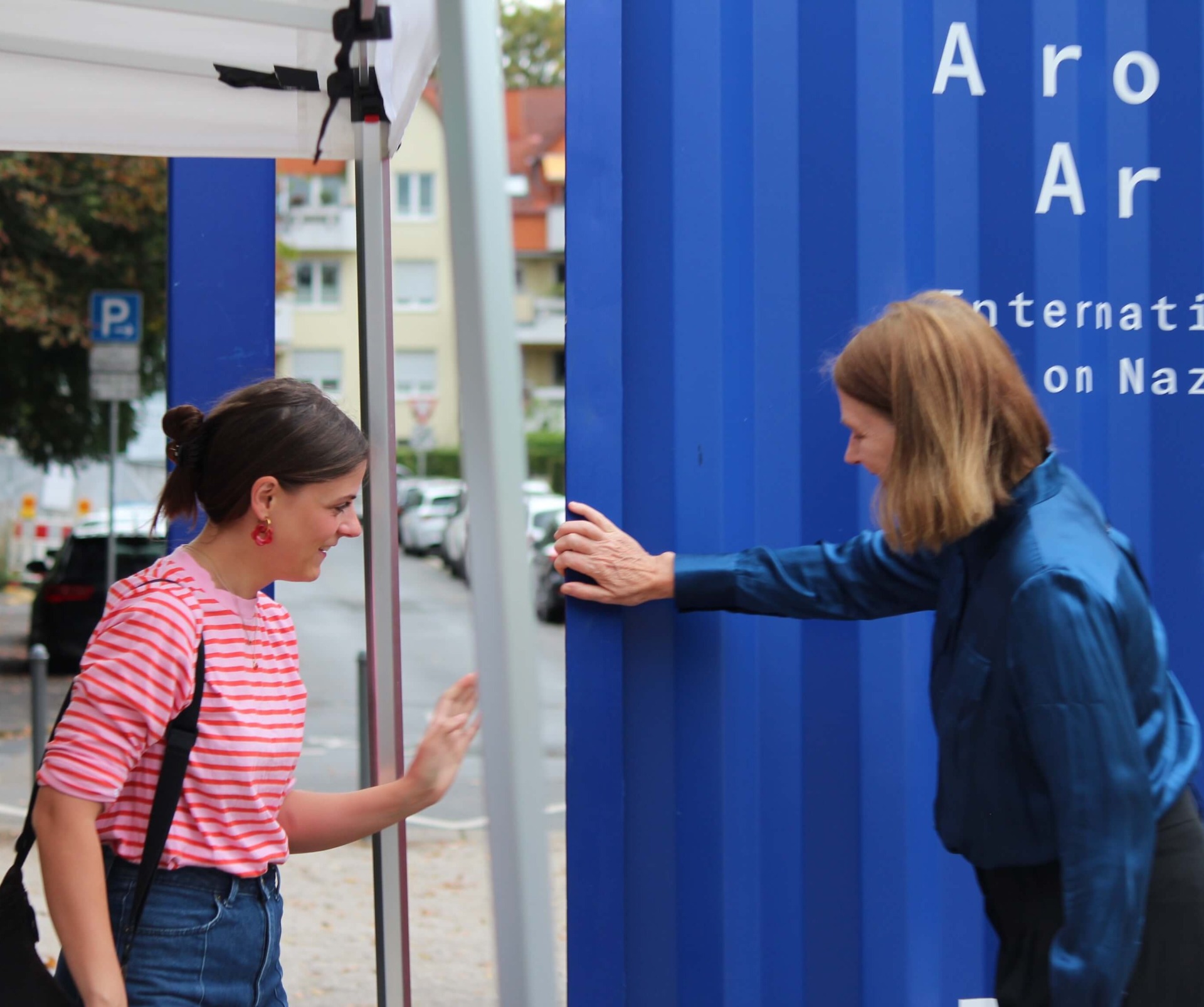 Zu sehen sind die Mitarbeiterin der Arolsen Archives Lilith Roska (links) und Bürgermeisterin Antje Runge während beide gemeinsam die Wandtür des Containers am Ausstellungsorts im Grünstreifen an der Adenauerallee öffnen.