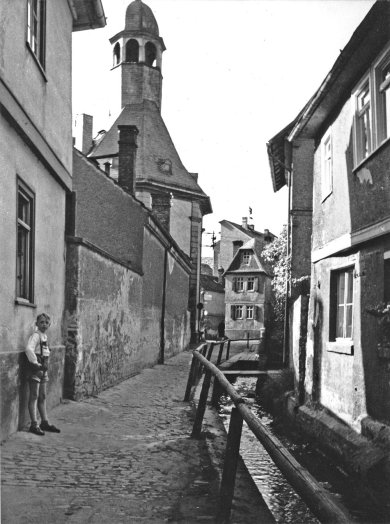 Man sieht die Hospitalkirche, davor eine geschlossene Mauer zum Platz und gegenüber den offenen Urselbachkanal, der dort entlang fließt wo heute der Eingang des Kulturcafes Windrose ist. 