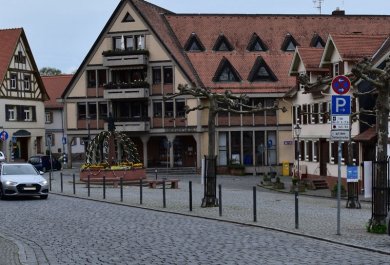 Gleicher Blickwinkel auf den Marktplatz wie auf dem alten Foto. Die alten großen Bäume sind weg. Es fahren viele Autos. Der Brunnen hat ein anderes Aussehen. Die Stadtbücherei steht anstelle eines großen alten Anwesens